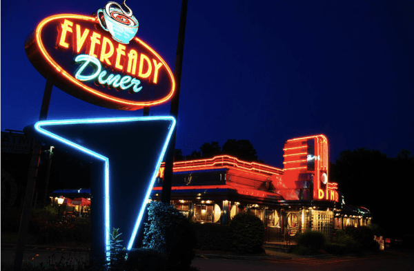 A brightly lit Eveready Diner sign glows in neon at night, as the classic diner building shines with red and orange neon lights against the deep blue sky.